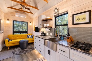 A modern kitchen with a yellow sofa, a dining table, and a large window. - La Petite: a Tiny House Tucked in Nature *Hot Tub* (Hays County)