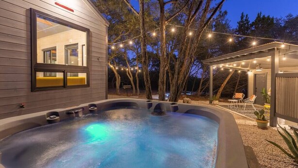 Un jacuzzi en un patio trasero con una casa de madera, luces colgantes y una zona de estar cubierta.