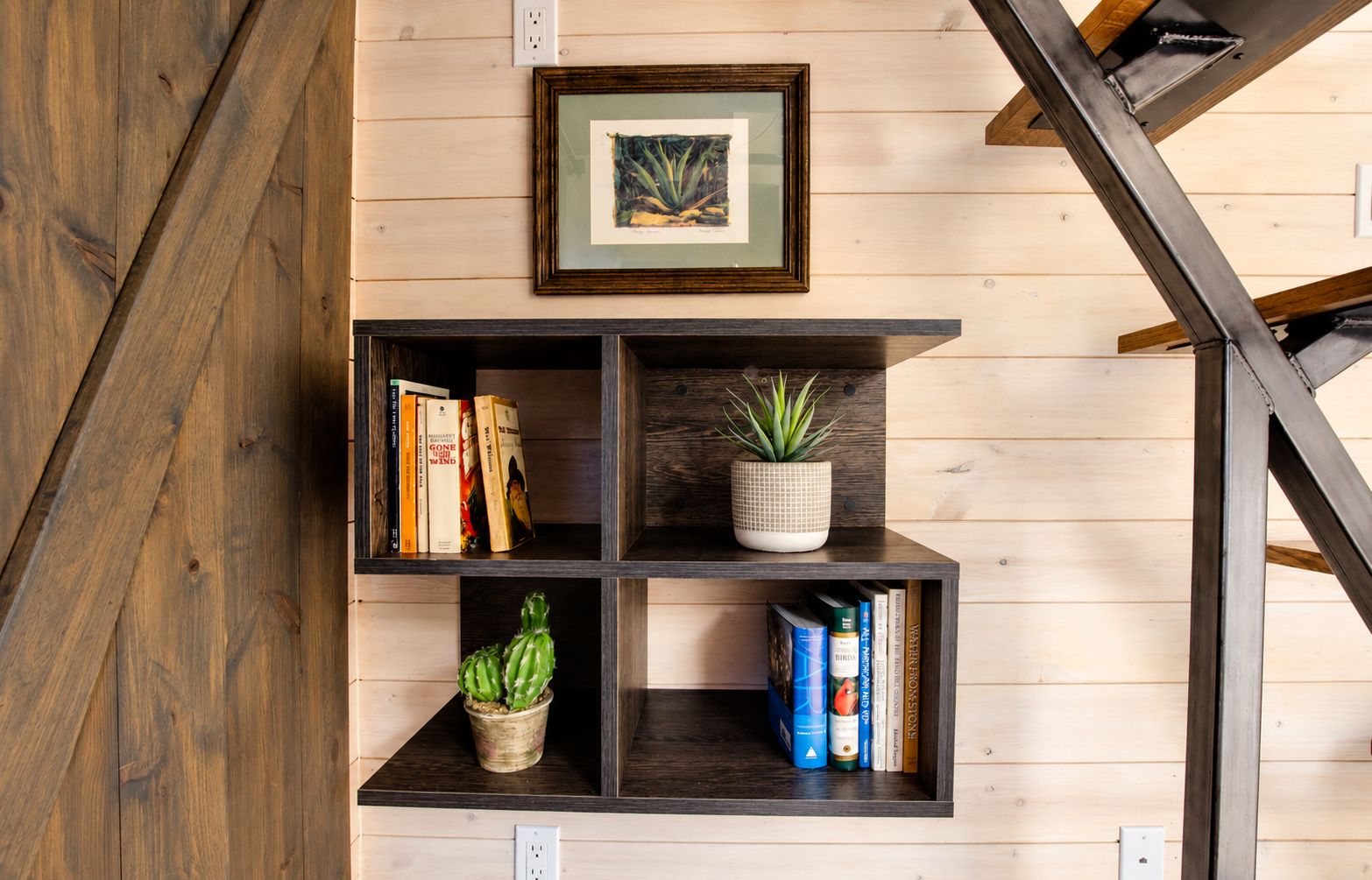 A wooden wall with a black metal staircase, a framed picture, and a shelf with books and plants.