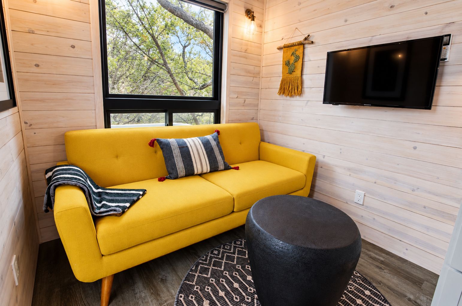 A yellow sofa with a patterned pillow, a black ottoman, and a mounted TV in a room with wooden walls and a window.