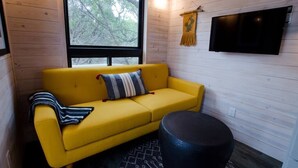 Un sofá amarillo con un cojín a rayas, un reposapiés redondo negro y un televisor montado en la pared.
