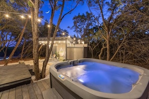 A hot tub surrounded by trees, a wooden deck, and a small building with string lights. - La Petite: a Tiny House Tucked in Nature *Hot Tub* (Hays County)