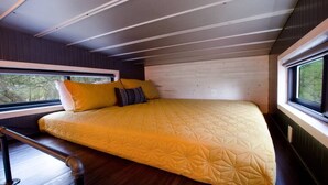 Una cama con colcha amarilla en una habitación tipo cabaña con paredes de madera y ventanas grandes.