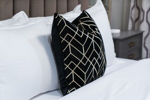 Una cama bien hecha con sábanas blancas y dos almohadas negras con diseños geométricos dorados.
