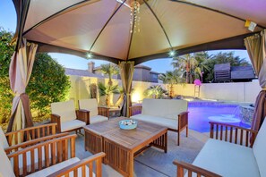 Un patio exterior con mesa y sillas de madera, una piscina y una zona cubierta con cortinas.