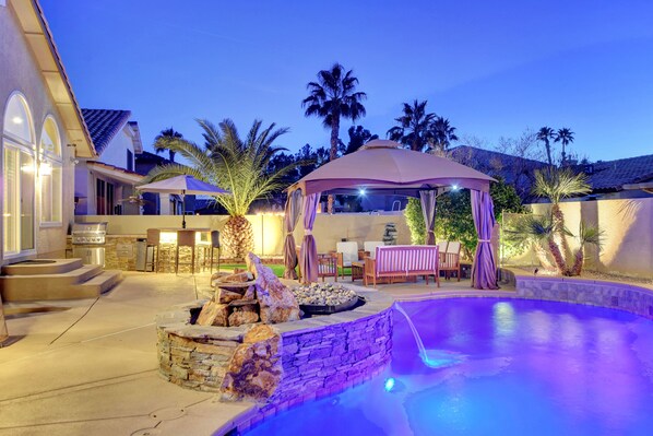 A backyard with a swimming pool, a stone fire pit, a covered patio, and a kitchen area.
