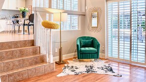 A modern living room with a staircase, a green armchair, a floor lamp, and a large window with blinds.