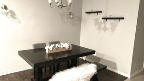 A dining area with a dark wooden table, a bench with a white furry seat, a chandelier, and two wall-mounted shelves. - Spacious Modern Retreat House (Garland)