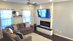 A living room with a sectional sofa, a flat-screen TV mounted on a stone fireplace, and a ceiling fan. - Spacious Modern Retreat House (Garland)