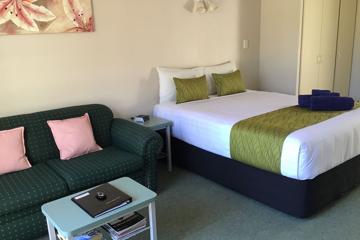 Ein Hotelzimmer mit einem grünen Sofa, einem Bett mit weißer und grüner Bettwäsche, einem kleinen Tisch mit einem Laptop und Büchern sowie einem Gemälde an der Wand.
