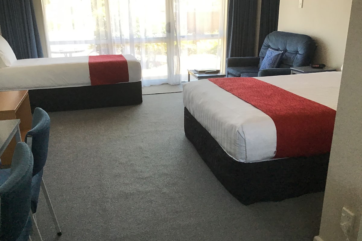 Ein Hotelzimmer mit zwei Betten, einem Schreibtisch, einem Sessel und Blick durch große Fenster ins Freie.