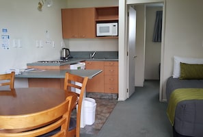 Una cocina compacta con armarios de madera, un microondas y un fregadero. Hay un comedor con una mesa y sillas. Se ve una cama en la habitación contigua.
