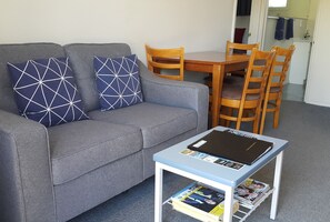 Un sofá gris con almohadones de estampado geométrico, una mesa de comedor de madera con cuatro sillas y una pequeña mesa blanca con revistas.
