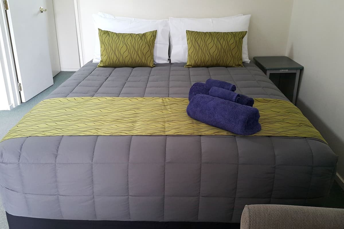 Ein Bett mit einer grauen Decke, zwei Kissen, einem gelb gemusterten Überwurf und zwei aufgerollten Handtüchern.