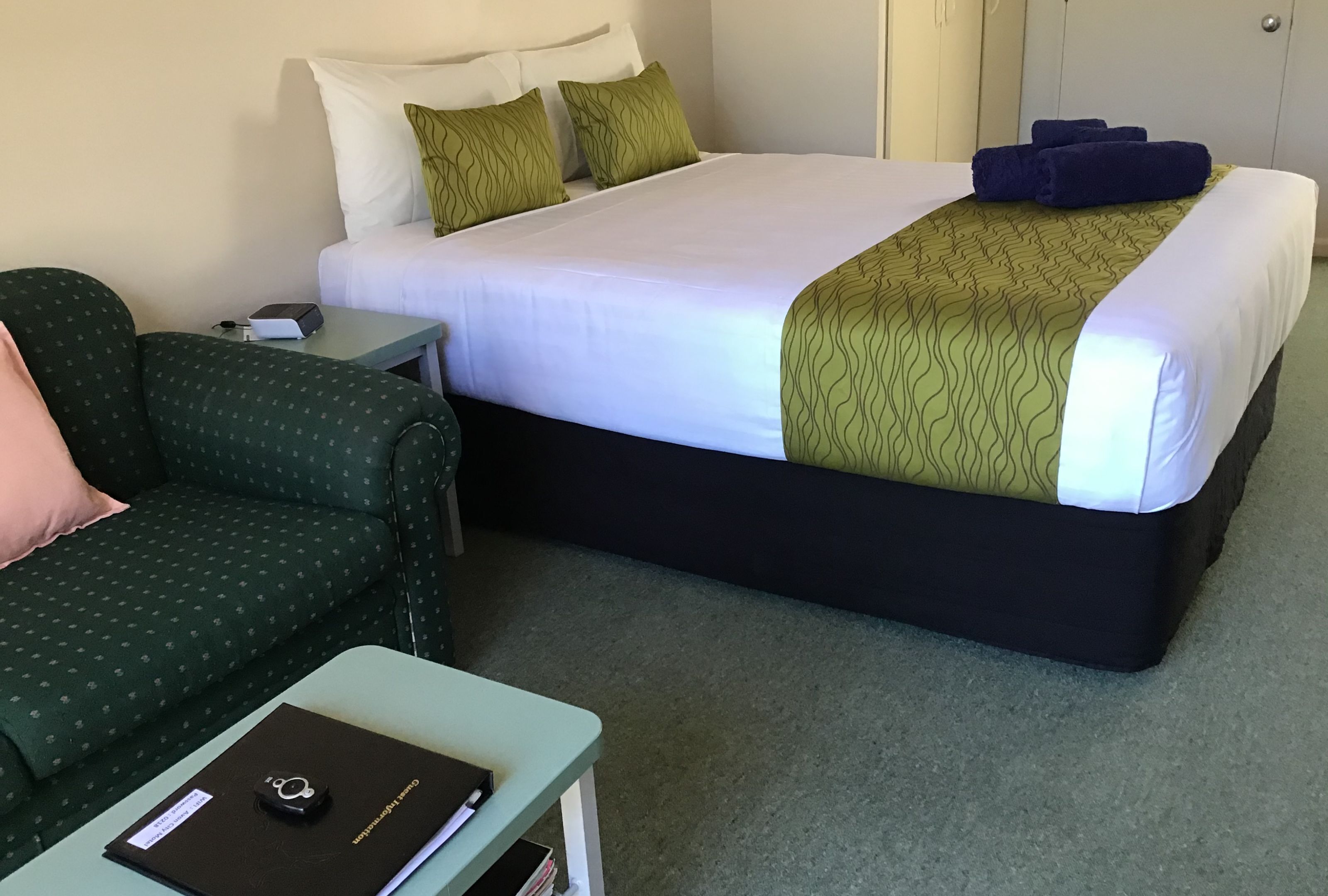Una habitación de hotel con una cama, un sofá verde estampado, una mesita con una carpeta y una mesita de noche.