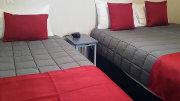 A hotel room with two beds, each with a grey quilted mattress and red pillows. There is a small table between the beds with a remote control on it.