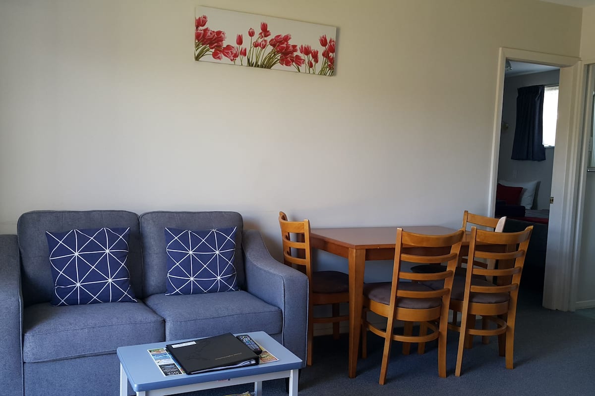Ein Wohnzimmer mit grauem Sofa, zwei gemusterten Kissen, einem kleinen Tisch mit Buch und Fernbedienung und einem Holzessttisch mit vier Stühlen.