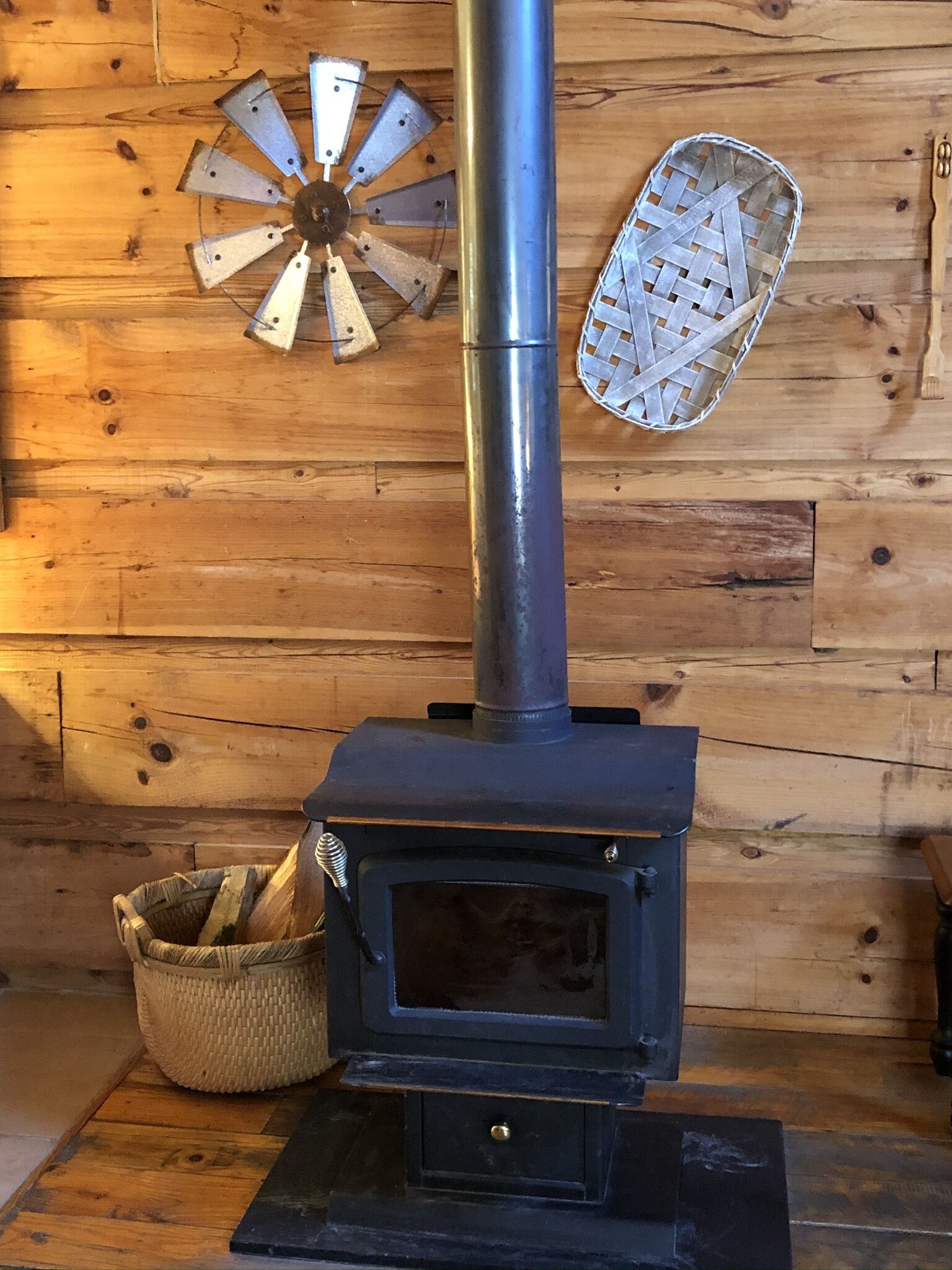 A wood stove with a metal chimney, a woven basket, and a woven wall decoration.