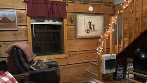 Ein gemütliches Blockhäuschen-Interieur mit Holzbalkendecke, einer schwarzen Ledersofa, einem Holztisch und einer Treppe, die zu einer oberen Ebene führt.