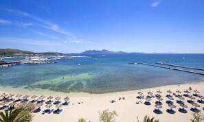 Ein Strand mit klarem Wasser, einer Marina und einem sandigen Ufer mit Sonnenschirmen und Liegestühlen.