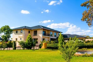 En bostadsbyggnad i två våningar med ett platt tak, flera balkonger och en välskött trädgård med en liten damm.