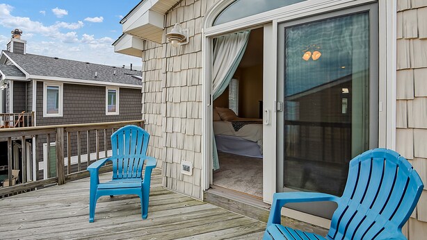 Een houten terras met twee blauwe stoelen, een schuifdeur naar een slaapkamer en een uitzicht op omliggende huizen.