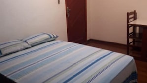 A bedroom with a bed, a wooden door, and a small table with a chair. - Chácara Paraíso (Miguel Pereira)