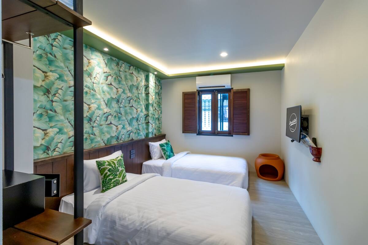 A hotel room with two beds, a wooden headboard, a floral wall design, a window with shutters, and a small orange planter.