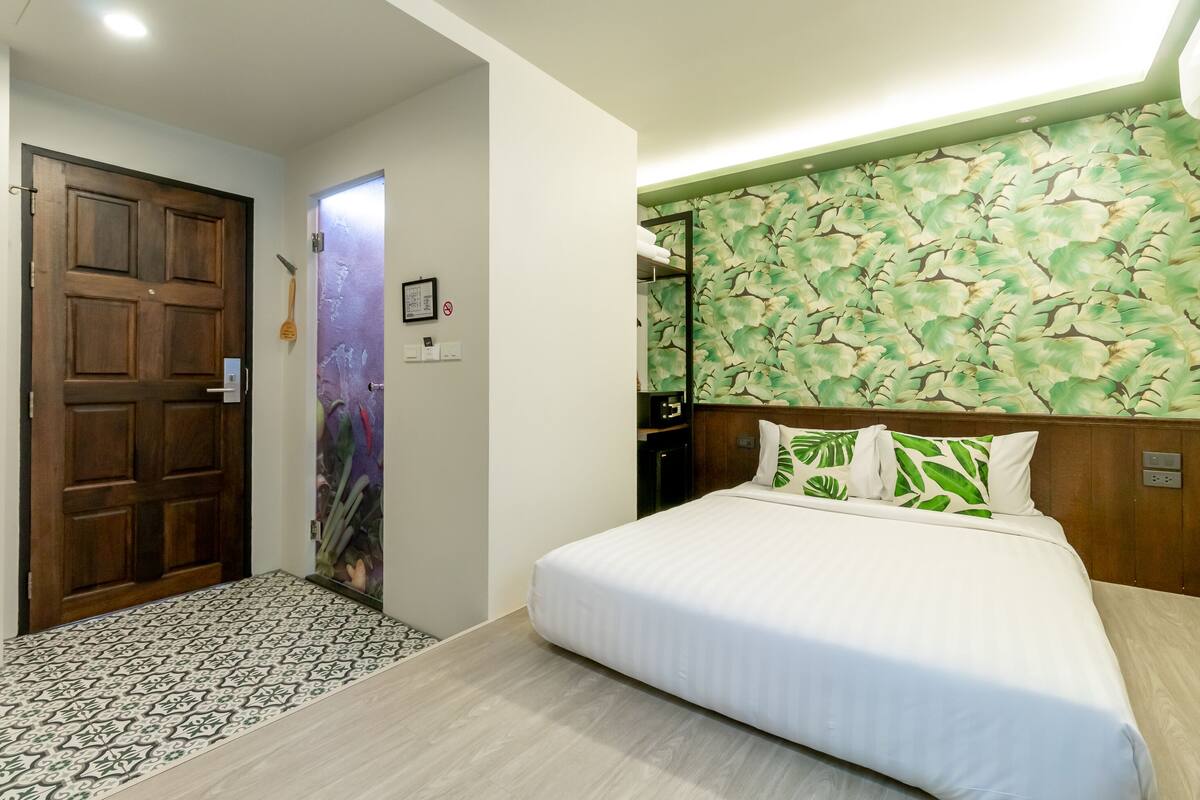 A hotel room with a wooden door, a patterned rug, a bed with white bedding, and a leaf-patterned headboard.