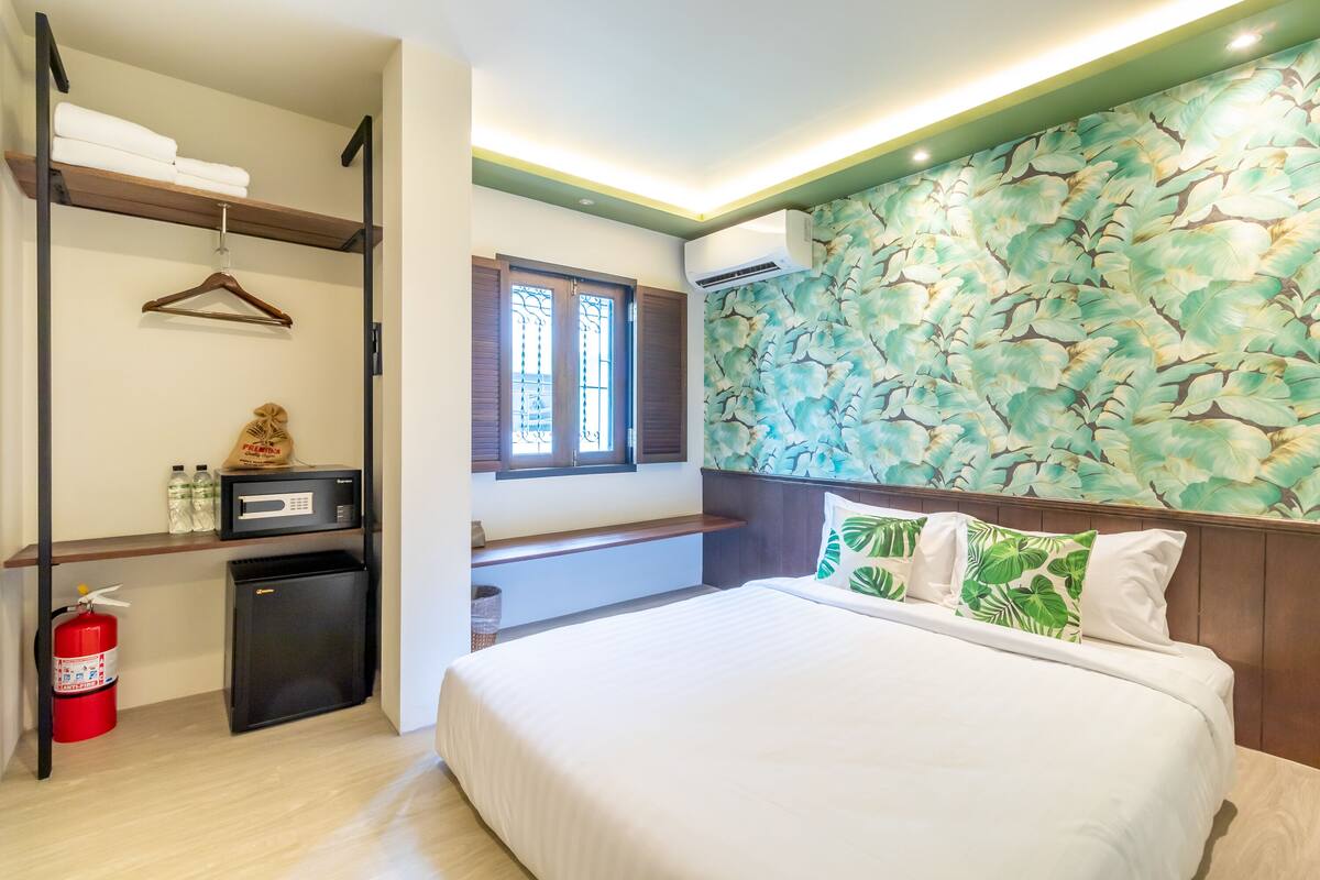 A hotel room with a bed, a small desk with a microwave, a fire extinguisher, and a window with shutters.