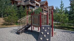 A playground with a climbing structure and a slide, surrounded by trees and a lodge in the background. - Spacious 3 BR Top Floor Condo - Kitchen, Mountain View Balcony, Fireplace & Pool (Canmore)