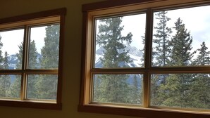 A room with large windows offering views of dense forest and snow-capped mountains. - Spacious 3 BR Top Floor Condo - Kitchen, Mountain View Balcony, Fireplace & Pool (Canmore)