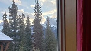 A window view of a mountainous landscape with tall evergreen trees and a wooden cabin structure. - Spacious 3 BR Top Floor Condo - Kitchen, Mountain View Balcony, Fireplace & Pool (Canmore)