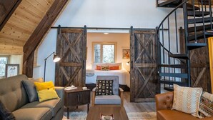 A loft-style room with a high ceiling, wooden beams, a staircase, a bed, a sofa, and a coffee table. - Salted Roots Red Door A-Frame Cabin (Seward)