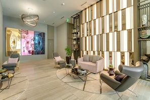 Ein modernes Wohnzimmer mit einem Glastisch, einer Couch und zwei Sesseln. An der Wand hängt ein großes abstraktes Gemälde und eine dekorative Ablagefläche mit Pflanzen.