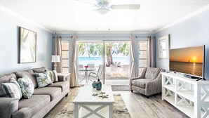 A living room with a sofa, armchair, coffee table, TV stand, and a view of the beach through the glass door. - White Sands 11-Beachfront Condo directly on 7 Mile Beach. Call Cayman Villas! (Seven Mile Beach)