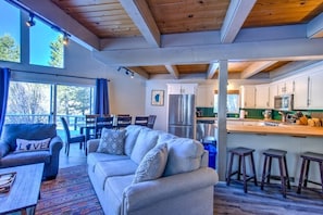 A modern living room with a sofa and a dining area, a kitchen with stainless steel appliances, and large windows offering a view of trees.