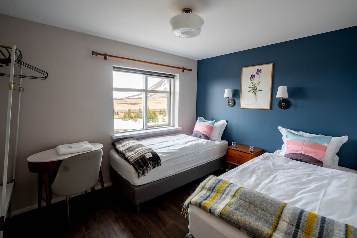 A bedroom with a bed, a small table, a chair, a window, and a wall-mounted lamp.