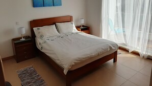 A neatly made bed with a white duvet, two bedside tables with lamps, a large window with sheer curtains, and a painting of a beach scene on the wall.