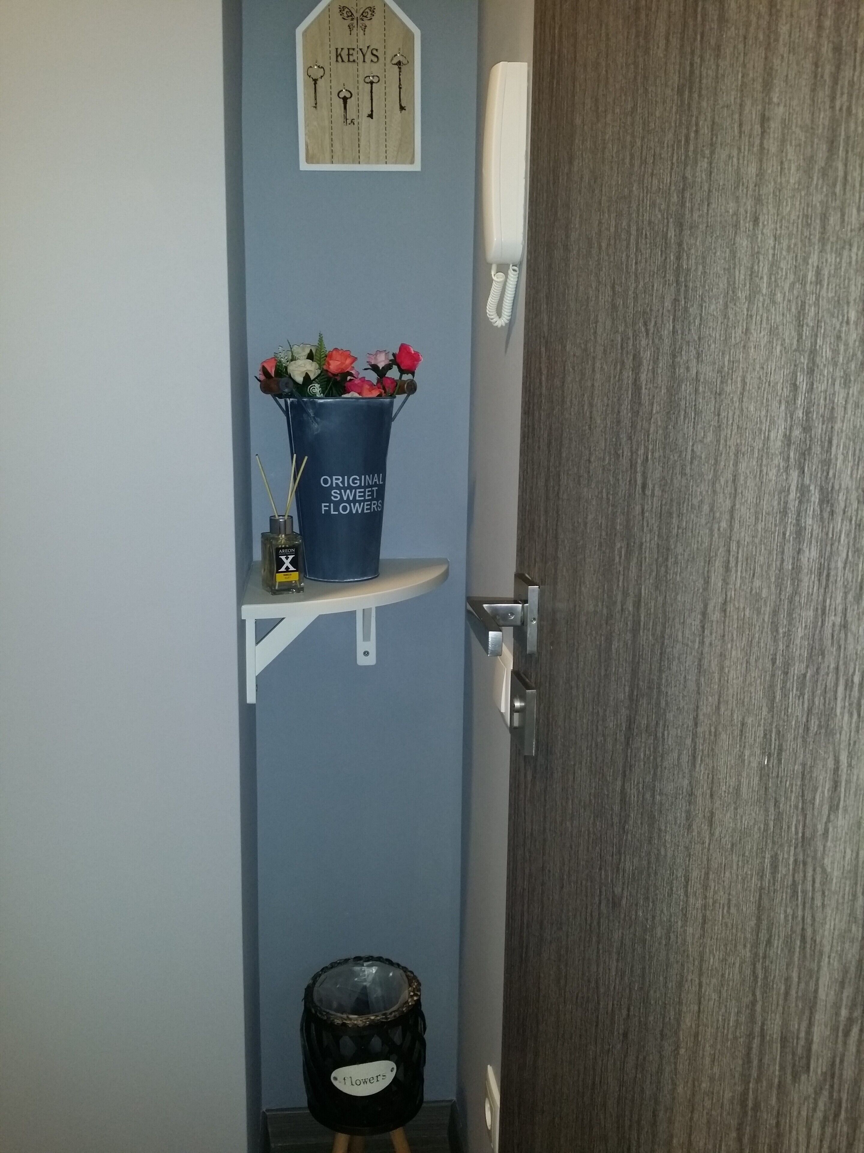 A bathroom with a blue wall, a wooden sign above the mirror, a white telephone, a blue bucket with flowers, a small trash bin, and a door with a silver handle.