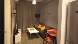 Een kleine woonruimte met een bank, een salontafel, een eettafel met stoelen, een plafondlamp en een koelkast.