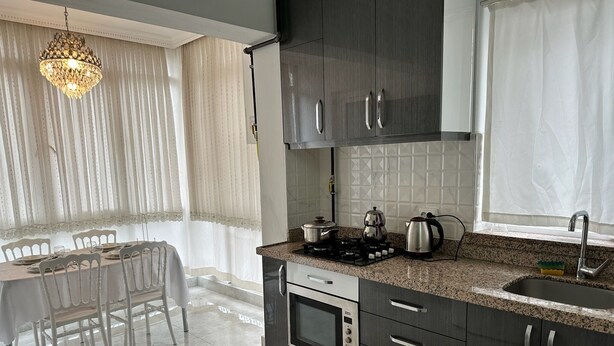 A modern kitchen with dark cabinets, a granite countertop, a built-in oven, and a window with sheer curtains. - Bulut Apartment (Konyaalti)