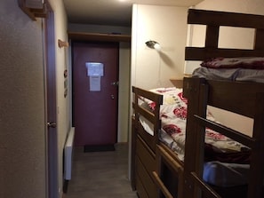 Un pasillo estrecho con una cama nido de madera a la derecha, una puerta al final y un escritorio pequeño con una lámpara.