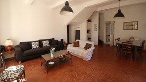 Amplia sala de estar con un sofá negro, un sofá blanco y una mesa de comedor con sillas. Hay lámparas colgantes, una mesa de centro y una lámpara. El piso está cubierto de baldosas y hay una escalera que lleva a otra habitación.