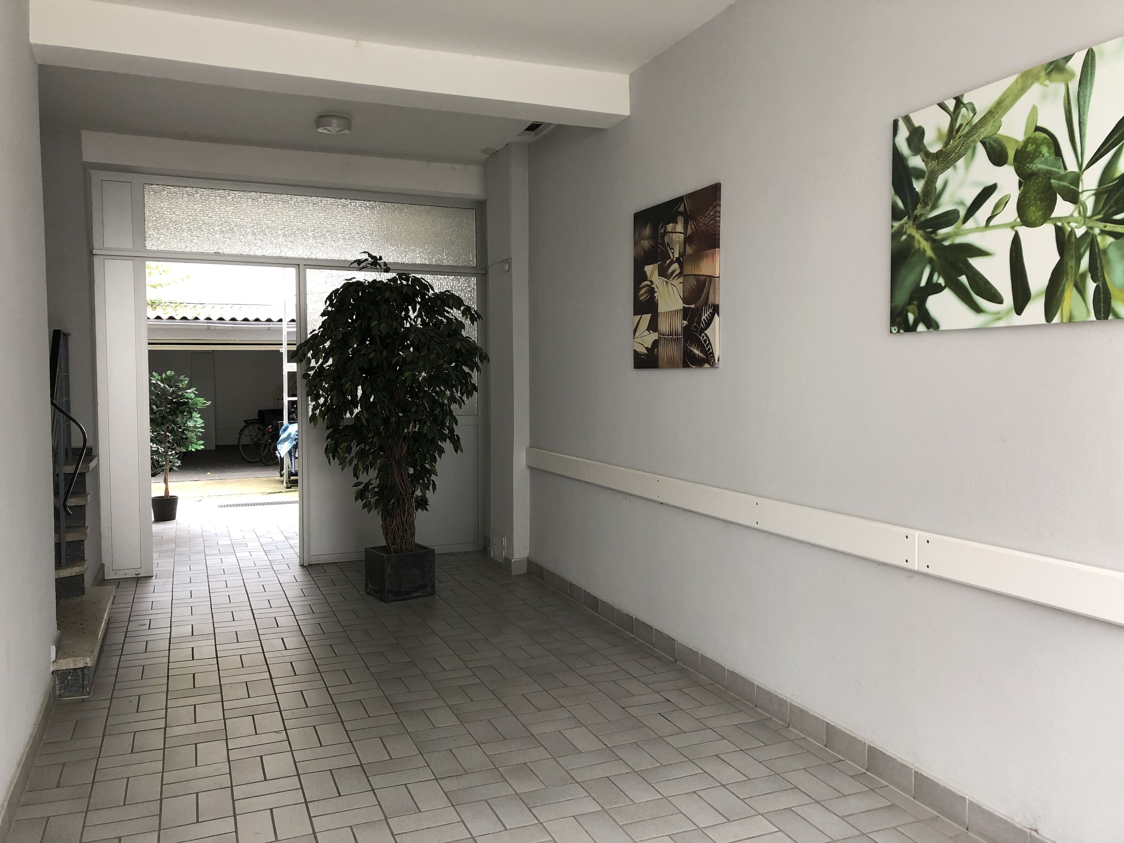 Een gang met een betegelde vloer, een potplant en twee kunstwerken aan de wanden.