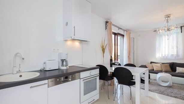 A modern kitchen with white cabinets, a built-in oven, and a coffee maker. There is a dining area with a white table and black chairs. - Villa Aurea (Čižići)