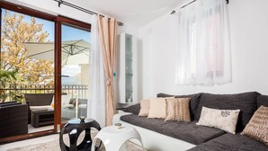 Een moderne woonkamer met een grijze bank, witte en gemêleerde kussens, een rond wit bijzettafeltje en een glazen deur die leidt naar een buitenruimte met rieten meubels en een boom.