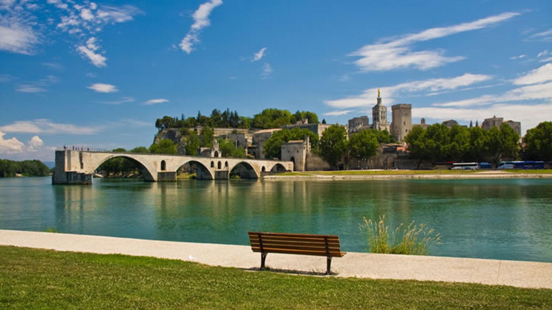 Une rivière traversée par un pont de pierre, avec un banc et des bâtiments historiques en arrière-plan.