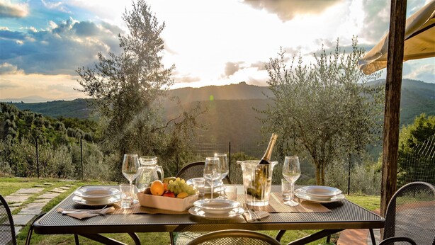 Eettafel buiten gedekt met borden, glazen en een fruitschaal, met uitzicht op een bergachtig landschap.