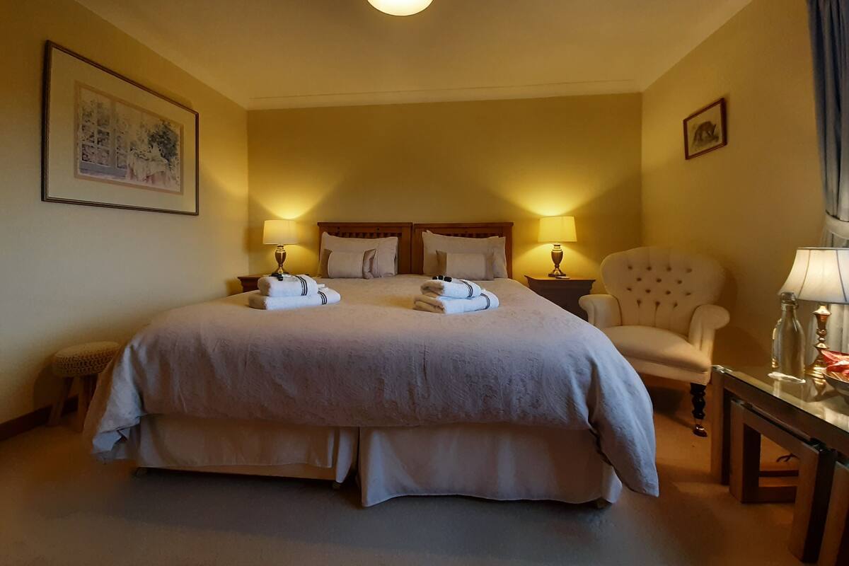A hotel room with a large bed, two bedside tables with lamps, a chair, and a small table with a bottle and glasses.
